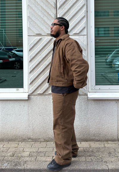 Chaqueta de algodón marrón y pantalones a juego, combinados con una camiseta negra. Gafas y cabello trenzado, de pie junto a una pared texturizada.