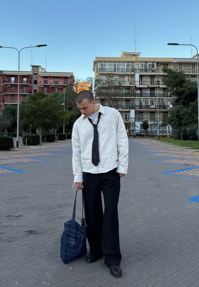 Camisa blanca de manga larga, corbata negra y pantalones anchos negros; sosteniendo una bolsa de mano de mezclilla azul. Colores sólidos, detalle mínimo, fondo urbano.