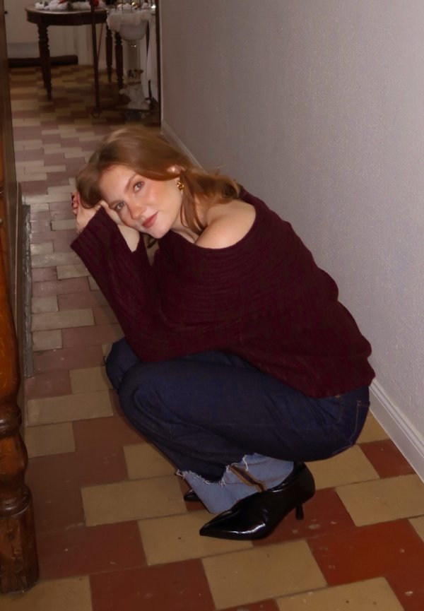 Maglione bordeaux lavorato a maglia grossa, jeans blu scuro con orlo sfrangiato e scarpe nere con tacco a punta. Persona accovacciata su pavimento piastrellato.