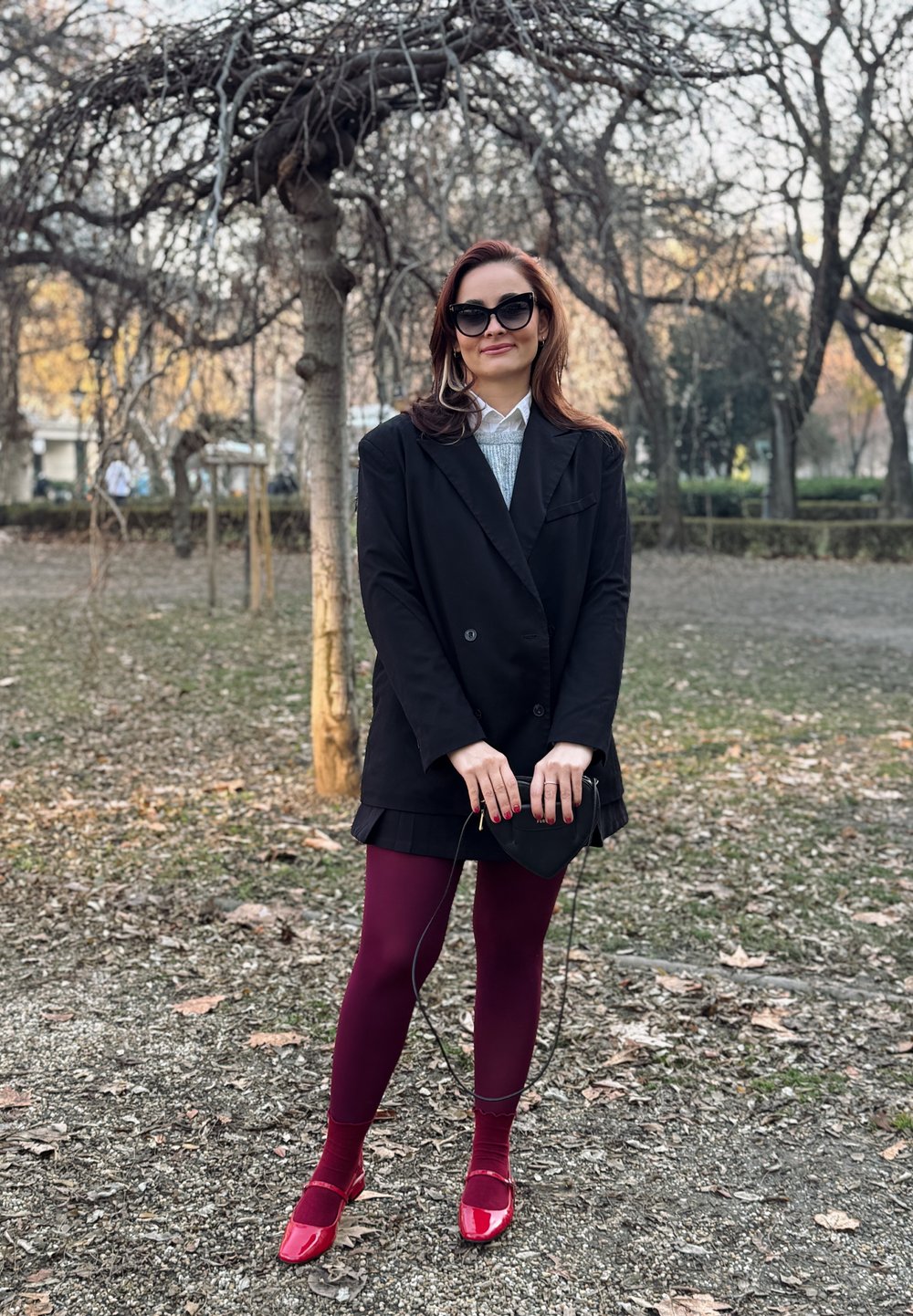 Μαύρο παλτό διπλής όψης πάνω από γκρι πλεκτό μπλουζάκι, μπορντό καλσόν και κόκκινα παπούτσια από λουστρίνι.  Κρατάει μια μικρή μαύρη τσάντα.