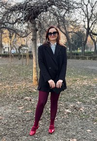 Μαύρο παλτό διπλής όψης πάνω από γκρι πλεκτό μπλουζάκι, μπορντό καλσόν και κόκκινα παπούτσια από λουστρίνι. Κρατάει μια μικρή μαύρη τσάντα.