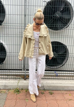 Chaqueta beige con cuello volado y botones frontales, llevada sobre una camiseta blanca sin mangas, combinada con pantalones anchos blancos y zapatos de color claro.