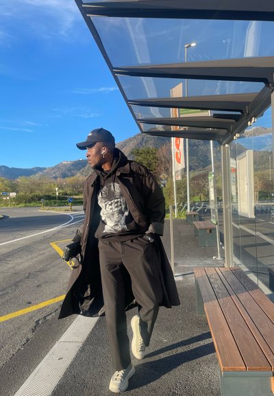 Abrigo negro sobre sudadera negra, pantalones negros y zapatillas de color claro. Los complementos incluyen un bolso negro y auriculares inalámbricos blancos.