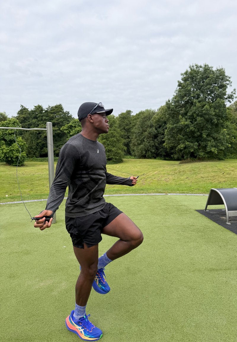 Man in sportswear skipping rope on a green outdoor exercise area with trees and grass in the background.