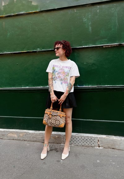 Mujer con cabello rizado, gafas de sol y tatuajes, vistiendo una camiseta gráfica, falda negra, tacones blancos, sosteniendo un bolso de mano con un patrón marrón y beige.