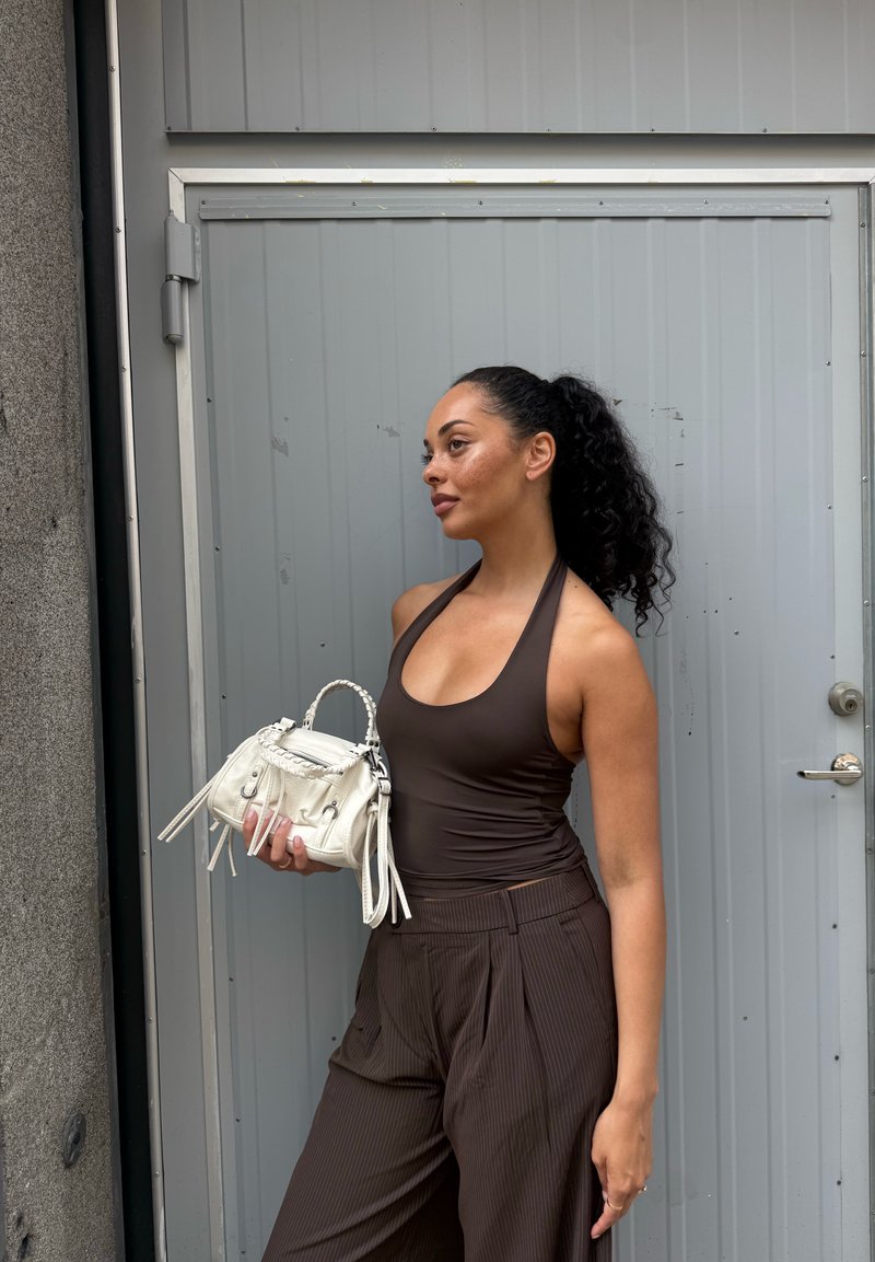 Femme aux cheveux bouclés portant un haut dos nu sombre et un pantalon à fines rayures, tenant un petit sac blanc, debout devant une porte métallique grise.