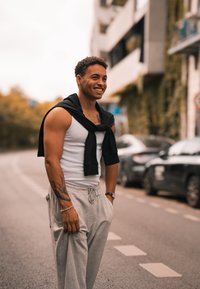 Λευκό αμάνικο μπλουζάκι και γκρι φόρμες, με ένα σκούρο πουλόβερ δεμένο γύρω από τους ώμους. Διαθέτει ορατό τατουάζ στο χέρι και ρολόι στον καρπό.