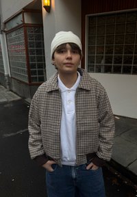 Veste bomber à carreaux marron et crème avec fermeture éclair, portée par-dessus un polo blanc. Un jean en denim de couleur foncée et un bonnet blanc complètent le look.