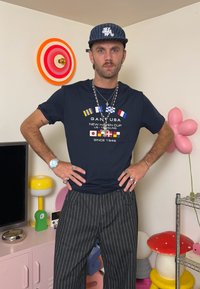 Tricou bleumarin cu steaguri de navigație colorate și text, asortat cu pantaloni gri cu dungi subțiri. Se poartă mai multe coliere și inele argintii.