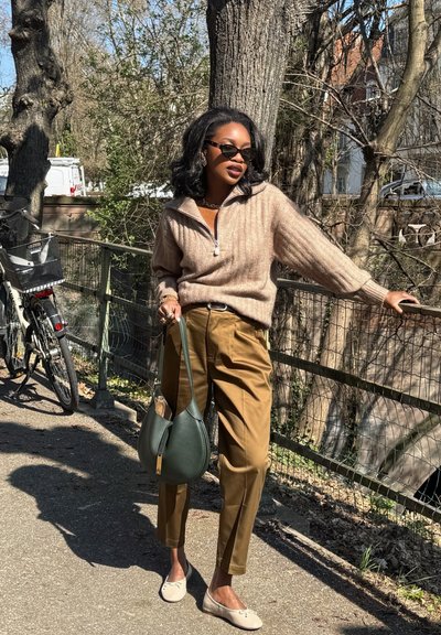 Suéter beige acanalado con cuello con cremallera; pantalones marrones a medida; bolso verde; zapatos planos de color claro; entorno al aire libre con árboles y bicicletas.