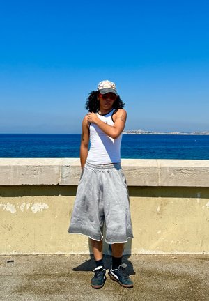 Persona con una camiseta sin mangas blanca y pantalones cortos grises de sudadera, con gafas de sol y una gorra con un diseño, de pie junto al océano con un cielo azul de fondo.