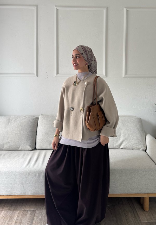 Femme portant une veste beige, un foulard à motifs, un pantalon large noir et portant un sac à main marron, debout dans un salon moderne.
