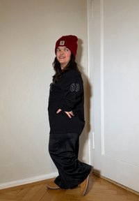 Czarna, oversize'owa bluza z detalem "69", zestawiona z szerokimi czarnymi spodniami i beżowymi butami. Na głowie czerwona czapka, stoi opierając się o ścianę.