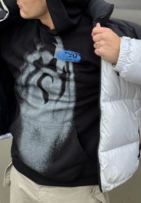 Zwarte hoodie met een grijs spattenpatroon en een blauw logo, gedragen onder een glanzend zilveren donzen jas. Lichtgekleurde broek zichtbaar.
