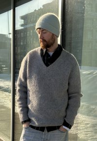 Homme portant un bonnet en laine gris, un pull gris à col en V sur une chemise à carreaux noire, et un jean bleu clair, debout à l'extérieur près de fenêtres en verre sous la lumière du soleil.