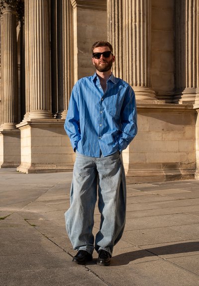 Hombre con gafas de sol negras, camisa azul con rayas y vaqueros anchos de color azul claro, de pie con las manos en los bolsillos frente a columnas de piedra.