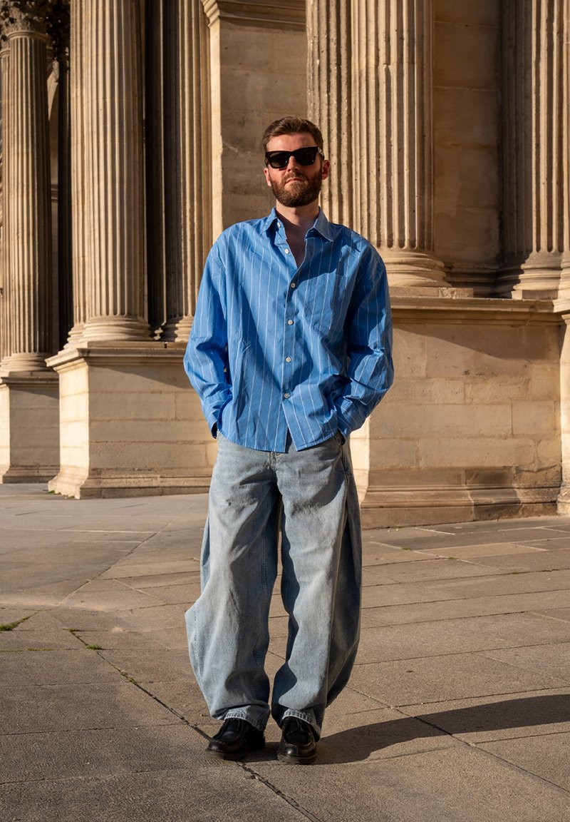 Homme portant des lunettes de soleil noires, une chemise bleue rayée et un jean large bleu clair, debout les mains dans les poches devant des colonnes en pierre.