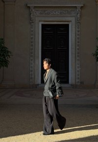 Mann, der im Sonnenlicht geht, trägt einen übergroßen dunklen Blazer und weite Hosen, hält eine schwarze Tasche und steht vor einer kunstvoll gestalteten großen dunklen Tür.