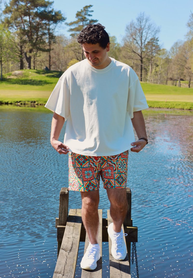 Homme portant un t-shirt blanc oversize et un short à motifs, marchant sur des planches de bois au-dessus d'un étang dans un parc ensoleillé.