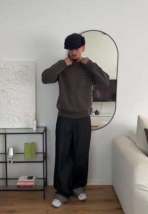 Suéter marrón oscuro con puños acanalados, presenta un pequeño logo. Pantalones de mezclilla negros holgados, zapatos blancos y una gorra negra. Espejo redondo en el fondo.