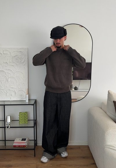 Suéter marrón oscuro con puños acanalados, presenta un pequeño logo. Pantalones de mezclilla negros holgados, zapatos blancos y una gorra negra. Espejo redondo en el fondo.