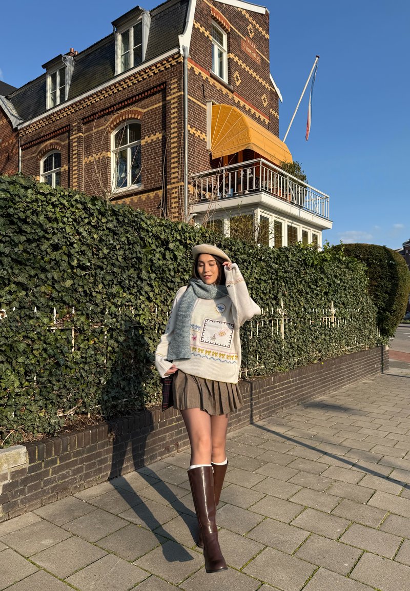 Fiatal nő bézs baretthel, túlméretezett pulóverrel, sállal, pliszírozott szoknyával és térdig érő csizmával áll egy járdán egy borostyánnal benőtt téglafal mellett.