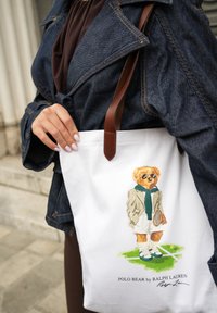 Balta rankinė su animaciniu lokiu, vilkinčiu švarką ir akinius nuo saulės, laikantis teniso raketę ir stovinčiu ant žalios žolės, su rudais odiniais diržais.