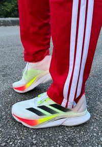 Ténis desportivos brancos com riscas pretas, apresentando uma sola em gradiente colorido e uma parte superior em malha leve, combinados com calças de fato de treino vermelhas.