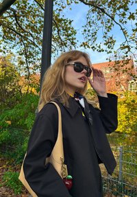 Veste noire à manches larges, sac à main beige avec un charme en forme de pomme, écharpe à motifs et lunettes de soleil sombres. L'arrière-plan présente de la verdure et un bâtiment.