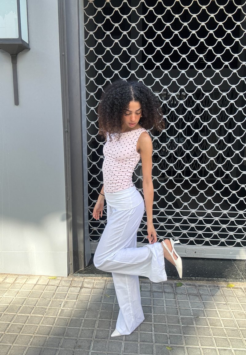 Femme aux cheveux bouclés touchant sa chaussure à talon blanc, debout sur un trottoir carrelé devant une grille métallique avec un motif incurvé.