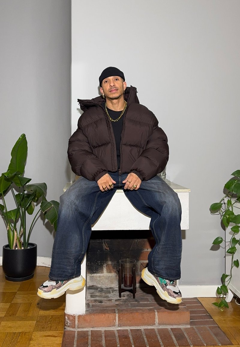 Giacca imbottita marrone scuro, maglietta nera, jeans blu oversize e scarpe da ginnastica multicolore. L'ambientazione include una pianta e un focolare in mattoni.