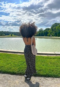 Eine Person steht an einem Teich und trägt ein schwarz-weißes Blumenkleid mit offenem Rücken sowie eine gemusterte Umhängetasche. Der Himmel ist bewölkt und es ist Grün in Sicht.