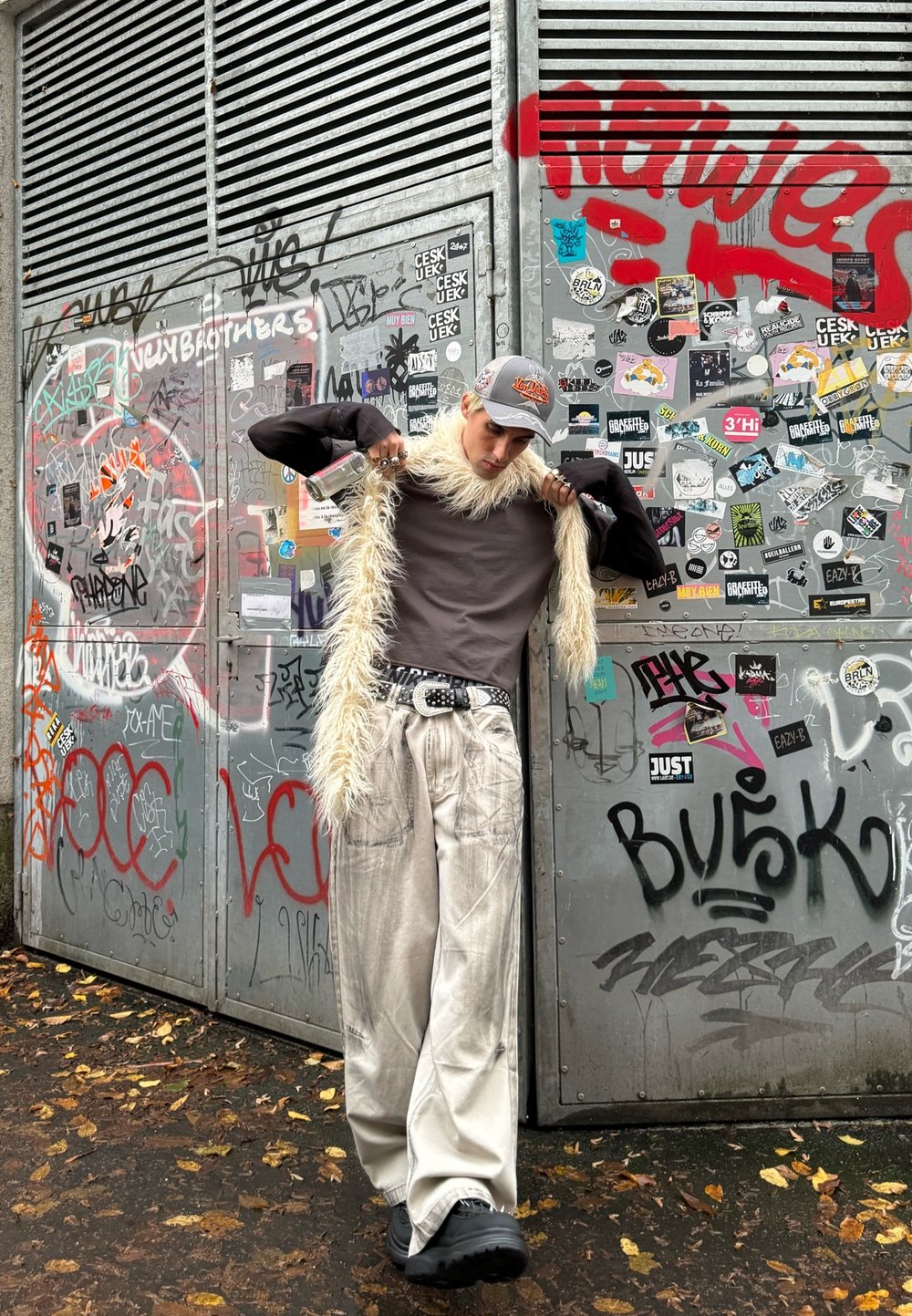 Model wearing a grey long-sleeve shirt, loose light grey trousers, and a fluffy white scarf. A cap with a logo and dark shoes complete the look. The background is covered in graffiti.