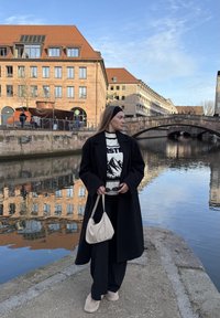 Manteau noir, pull blanc à motifs avec design de montagne, pantalon noir, sac à main beige et bottes de couleur claire. Rivière et bâtiments en arrière-plan.