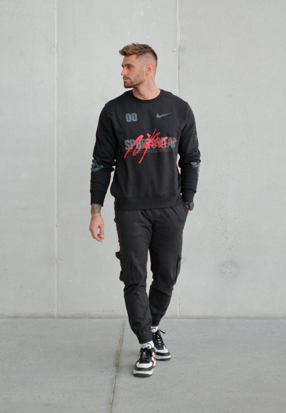 Sweat-shirt noir avec des graphiques rouges et gris, assorti à un pantalon cargo noir et des baskets noires. Le modèle se tient devant un mur gris.