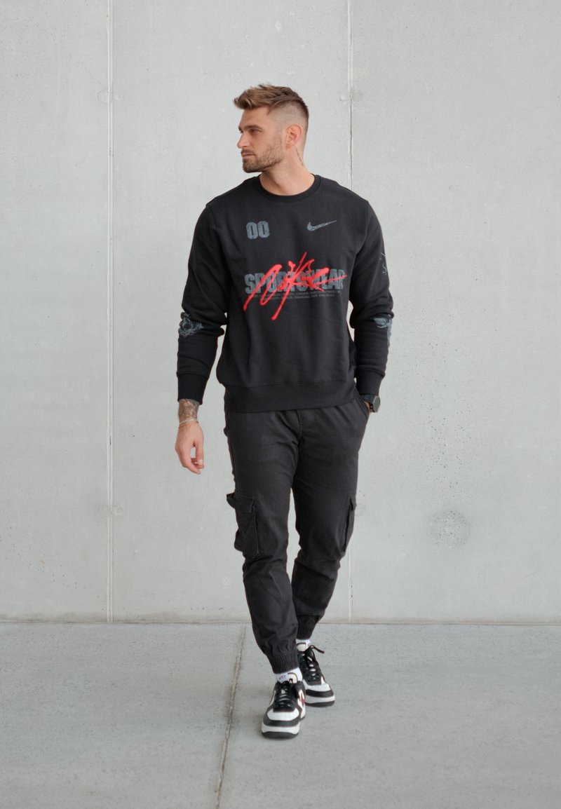 Sweat-shirt noir avec des graphiques rouges et gris, assorti à un pantalon cargo noir et des baskets noires. Le modèle se tient devant un mur gris.