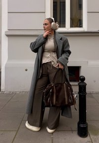 Grijze overjas, beige top met strik, bruine wijde broekspijpen, witte schoenen en een bruine schoudertas met ritsen. Neutrale stedelijke achtergrond.