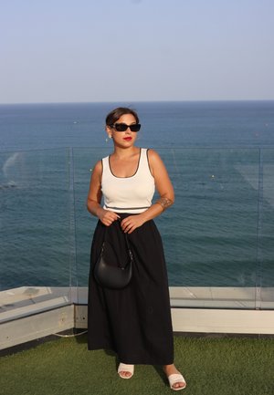 Mujer vestida con una camiseta sin mangas blanca, falda larga negra, gafas de sol y sosteniendo un bolso negro, de pie junto a una barandilla de vidrio con fondo oceánico.