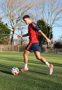 Jeune homme en tenue de football rouge et bleue lançant un ballon de football sur un terrain vert avec des arbres sans feuilles en arrière-plan sous un ciel clair.
