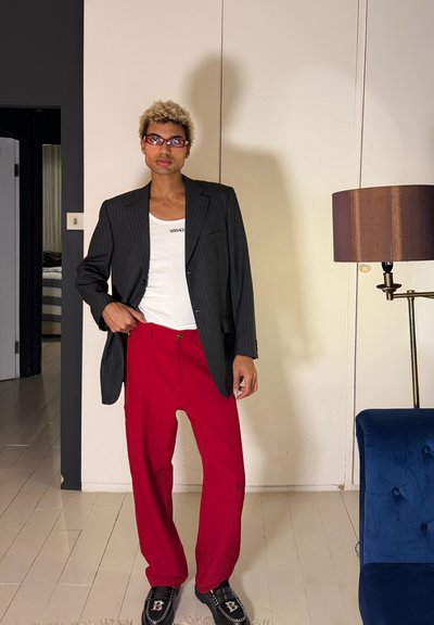 Persona joven con cabello rubio corto y rizado, vistiendo pantalones rojos, blazer negro a rayas finas, camiseta de tirantes blanca de Versace, gafas de sol rojas, de pie en interiores.