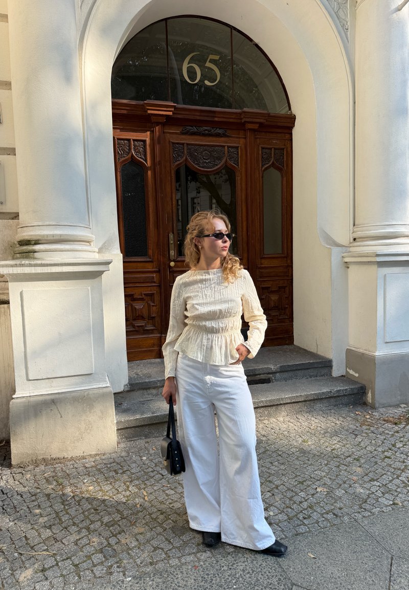 Woman wearing sunglasses, cream blouse, and white pants stands holding black bag outside a building with wooden double doors numbered 65.