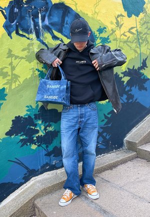 Jeans de mezclilla azules, chaqueta de cuero negra, sudadera con capucha negra con el texto "OFF LIMITS", y zapatillas deportivas naranjas. Sosteniendo una bolsa SAMSØE azul con texto blanco.