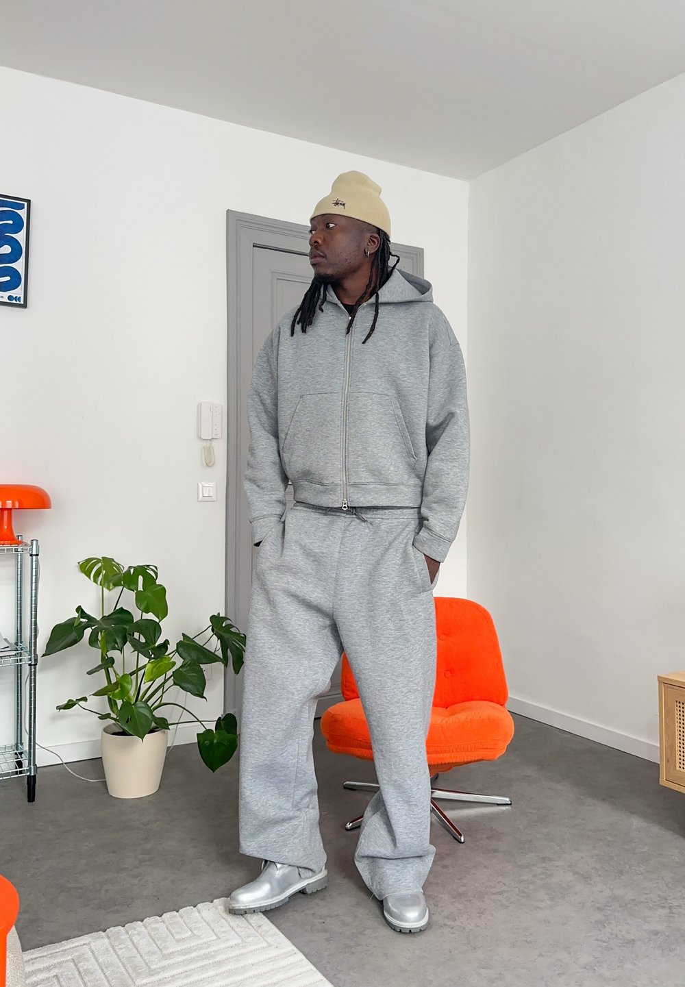 Mand iført grå hættetrøje, matchende joggingbukser, beige hue og sølvfarvede støvler står i et moderne rum med orange stol og grøn plante.