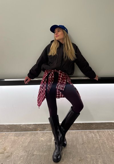 Joven mujer con chaqueta negra, camisa de cuadros roja atada a la cintura, leggings negros, botas altas negras y gorra azul apoyada contra una pared en el interior.
