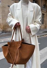 Manteau en laine crème sur un pull côtelé beige, tenant un sac fourre-tout en daim marron avec deux longues bandoulières, sur fond de rue.