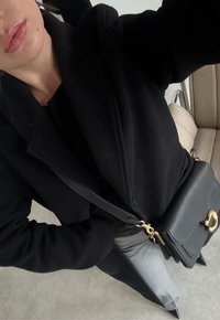 Femme portant un manteau noir, un pantalon gris et des chaussures pointues noires, portant un sac bandoulière en cuir noir avec des ferrures dorées.