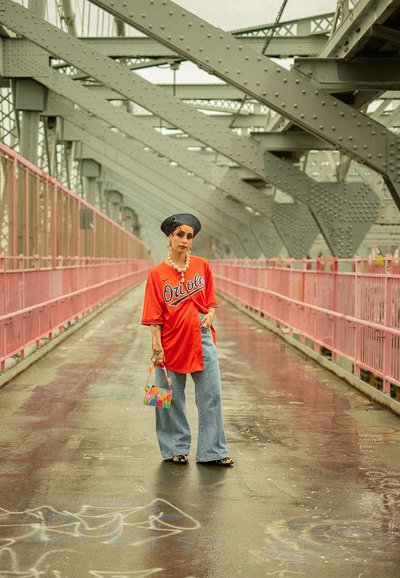 Camiseta oversized naranja con el texto "Orioles", jeans de pierna ancha en azul claro y un bolso tejido colorido, todo dispuesto sobre un puente rosa.