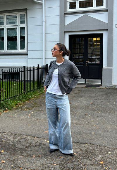 Cárdigan gris de punto, camiseta blanca y vaqueros de pierna ancha en azul claro. Accesorios con gafas negras. Entorno al aire libre con un fondo de edificio.