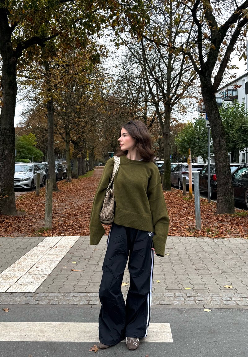 Femme portant un pull vert olive et un pantalon de jogging noir, debout sur un passage pour piétons dans une rue couverte de feuilles, bordée d'arbres d'automne dénudés et de voitures garées.