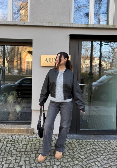 Chaqueta de cuero negra corta sobre un suéter gris, combinada con unos vaqueros grises de pierna acampanada y botas marrones. Sosteniendo un pequeño bolso negro. Entorno urbano moderno.